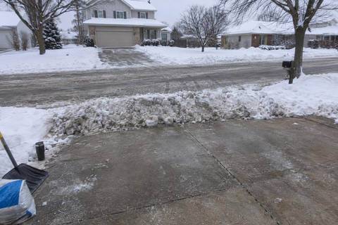 A ridge of dense plowed snow lying across the end of a concrete driveway where it meets a plowed suburban street, tire tracks in the road beyond it