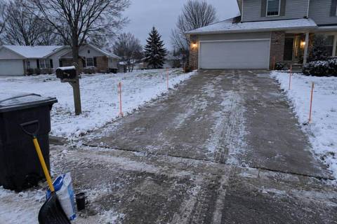 Orange driveway marker stakes lining both edges of a long asphalt driveway under fresh snow, a trash can and shovel at the near end and a two-story house with an attached garage at the far end