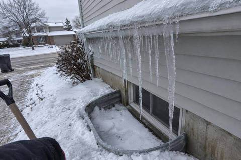 Thick icicles along a gutter edge above a basement window well filled with snow at the foundation of a vinyl-sided house