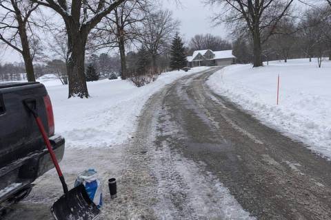 A long private driveway curving between bare oaks on rolling ground, one plowed lane down the center and deep snow on both shoulders
