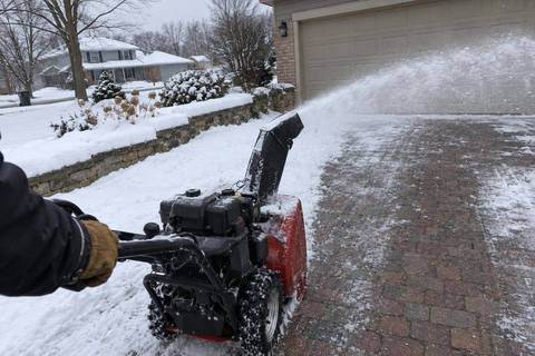 A snowblower working across a brick paver driveway, the paver pattern showing through the cleared strip and snow piled along a low garden wall