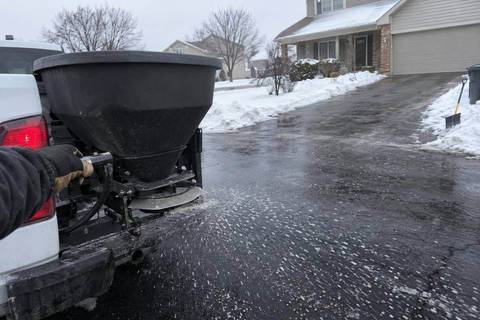A tailgate spreader on the back of a pickup throwing granular ice melt across a wet asphalt driveway, pellets scattered on dark pavement