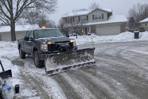 A pickup with a straight plow blade angled in a snow-covered cul-de-sac, plowed banks along the curb and a two-story house with an attached garage behind