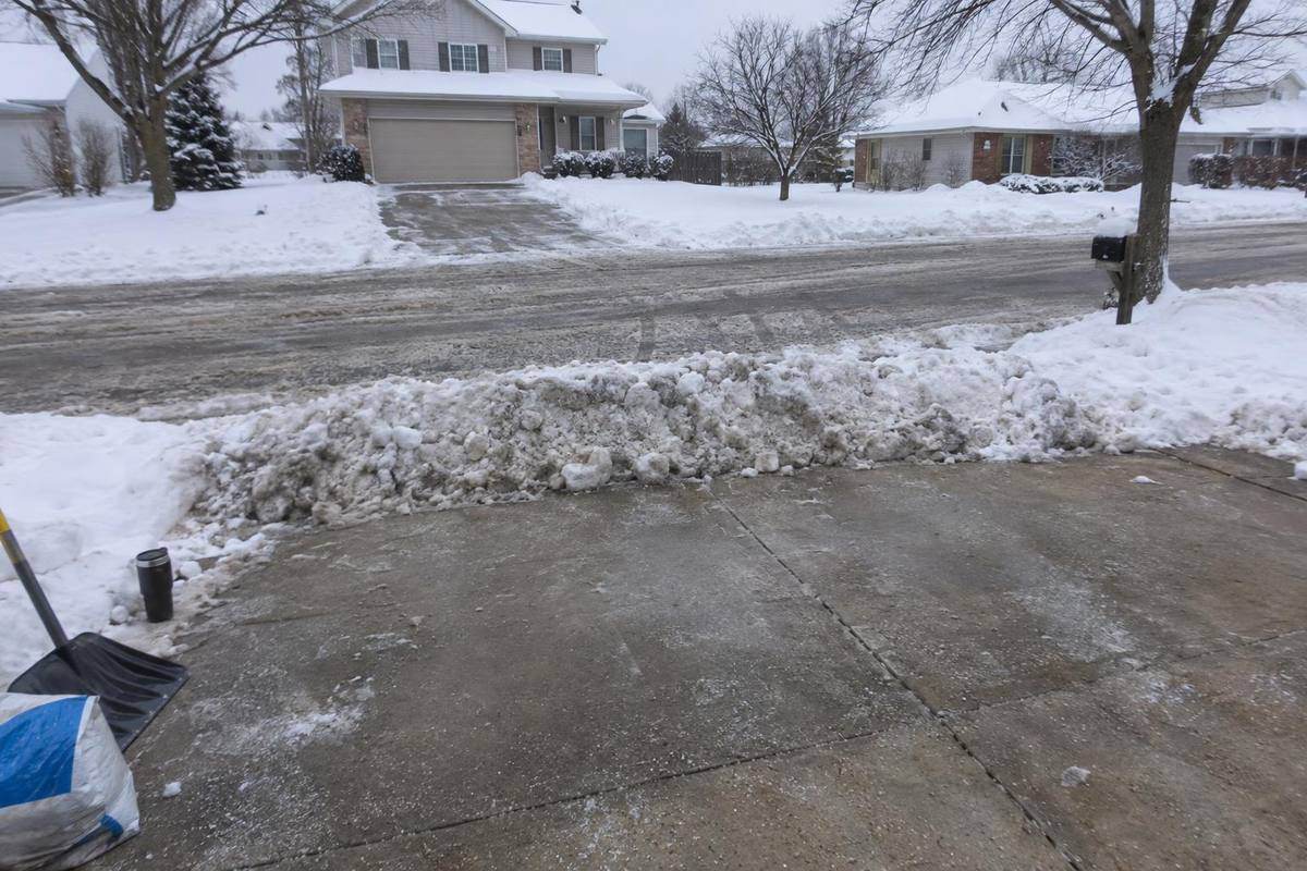 A dense ridge of plowed snow lying across the end of a concrete driveway where it meets a plowed street, with a mailbox post at one side