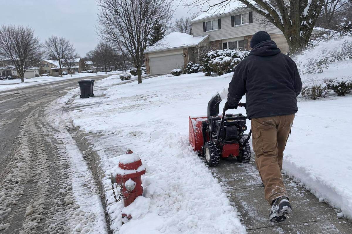 A walk-behind snowblower on a corner sidewalk with a curb ramp, snow banked on the lawn side and a street sign in the background